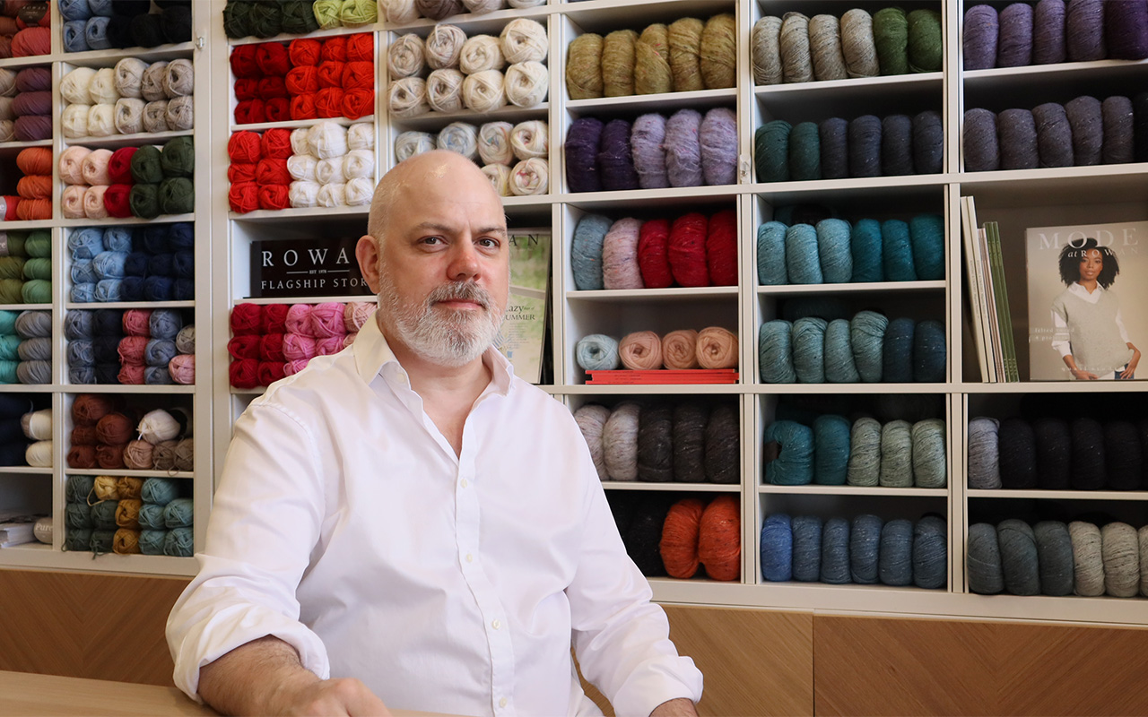 Stephen sitting in The Orry Mill wool shop infront of shelves of colourful yarn
